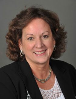 Headshot of WVU professor Nany McIntyre. She is middle-aged with brown, shoulder-length curly hair. She is wearing a black jacket over a white, lace top. She has a silver necklace and earrings on. 
