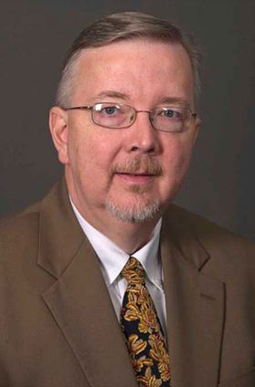 Headshot of retired WVU employee Les Carpenter. He is pictured in front of a dark taupe background wearing a brown coat over a white dress shirt with a black and yellow floral tie. He has short graying brown hair and glasses. 