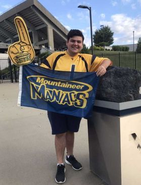 Man with dark hair wearing gold WVU jersey, holding up a blue flag that says "Mountaineer Maniacs," standing in front of a football stadium.
