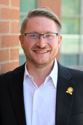 Headshot of WVU professor Sam Taylor. He is pictured outside in front of a red brick building. He is wearing a dark colored jacket with a gold lapel pin over a white dress shirt. He has short dark, blonde hair and a 5 o'clock shadow. He also wears glasses