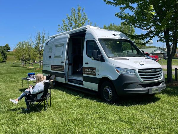 Photo of the Westbrook Health Services Mobile Unit, a converted cargo van, parked in a grassy area with support staff sitting nearby. 