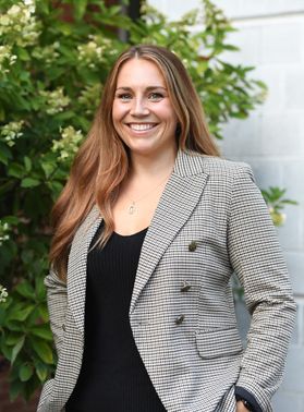 WVU researcher Erienne Olesh. She is pictured outside wearing a gray plaid jacket over a black blouse. She has long, light brown hair and has her hands in her pockets. 