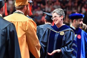 Provost Maryanne Reed, dressed in Commencement regalia, smiles while shaking the hand of a graduate wearing a gold cap and gown.