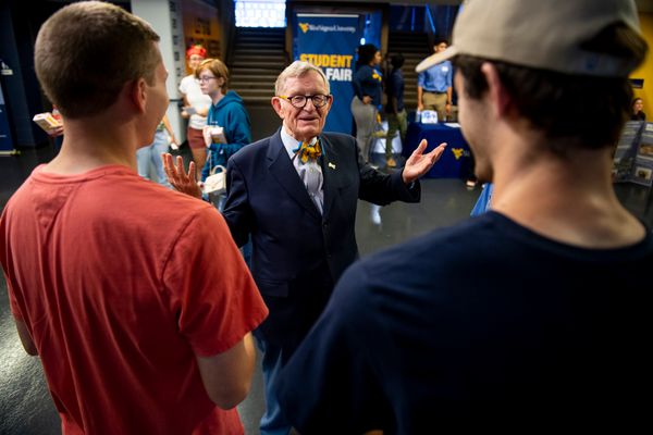 WVU President Gordon Gee stands in the middle of this photo. He is facing the camera while wearing a dark suit and bow tie. His left hand is raised. Two people listening have their backs to the camera. One is in a short-sleeved red shirt.