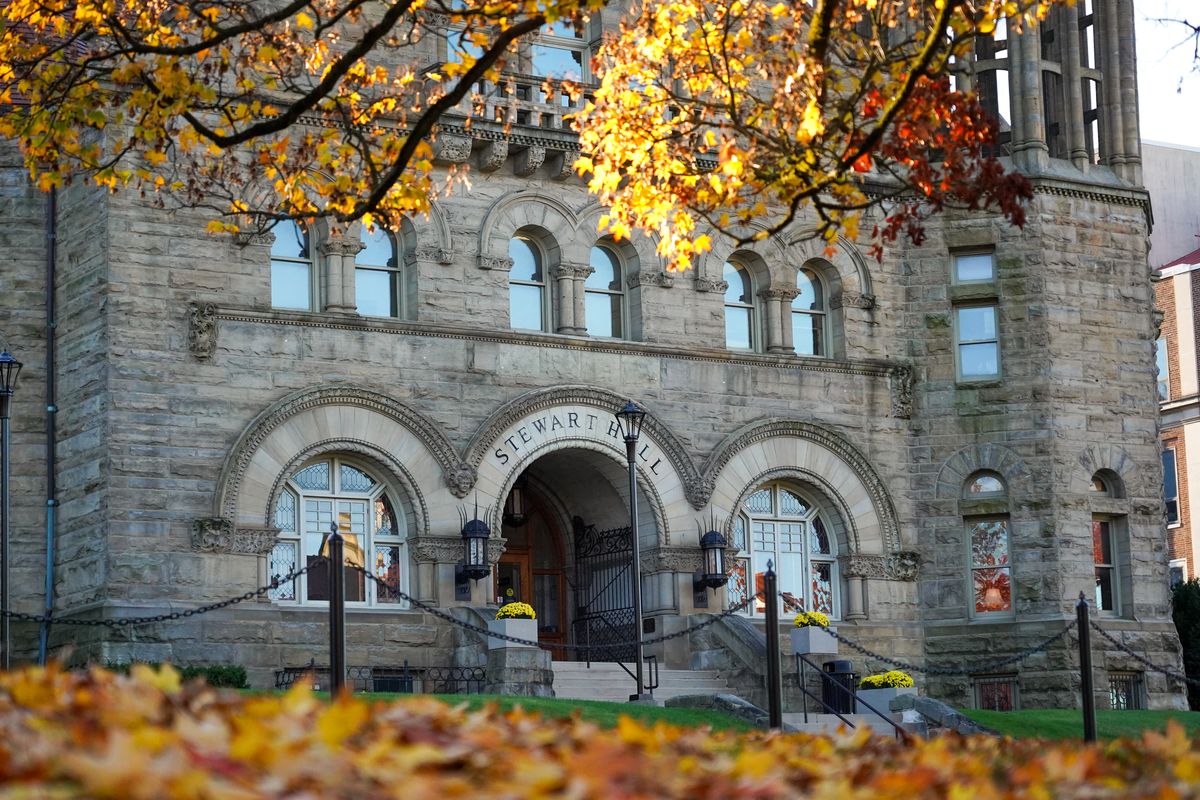 Photograph of Stewart Hall on the WVU Morgantown Campus in the fall. There are fallen leaves of orange and gold on the ground and still a few hanging in the trees in front of the gray brick building. 