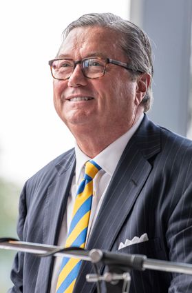 This is a portrait of Bob Reynolds who is wearing a dark striped suit jacket, a gold and blue tie and glasses.