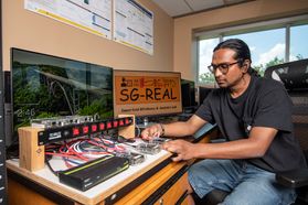 : Mohammed Mustafa Hussain, a computer science graduate student, is shown here working with equipment in the lab. He has long dark hair pulled back in a ponytail, a dark colored shirt and blue jeans. He is also wearing glasses. 