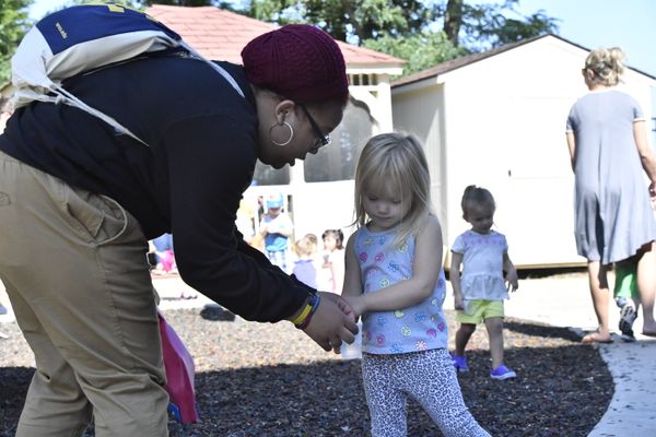 An image showing children playing on a playground. A teacher, wearing khaki pants, a dark colored shirt, a maroon hat and a blue and white backpack, is helping a young girl with blonde hair. 