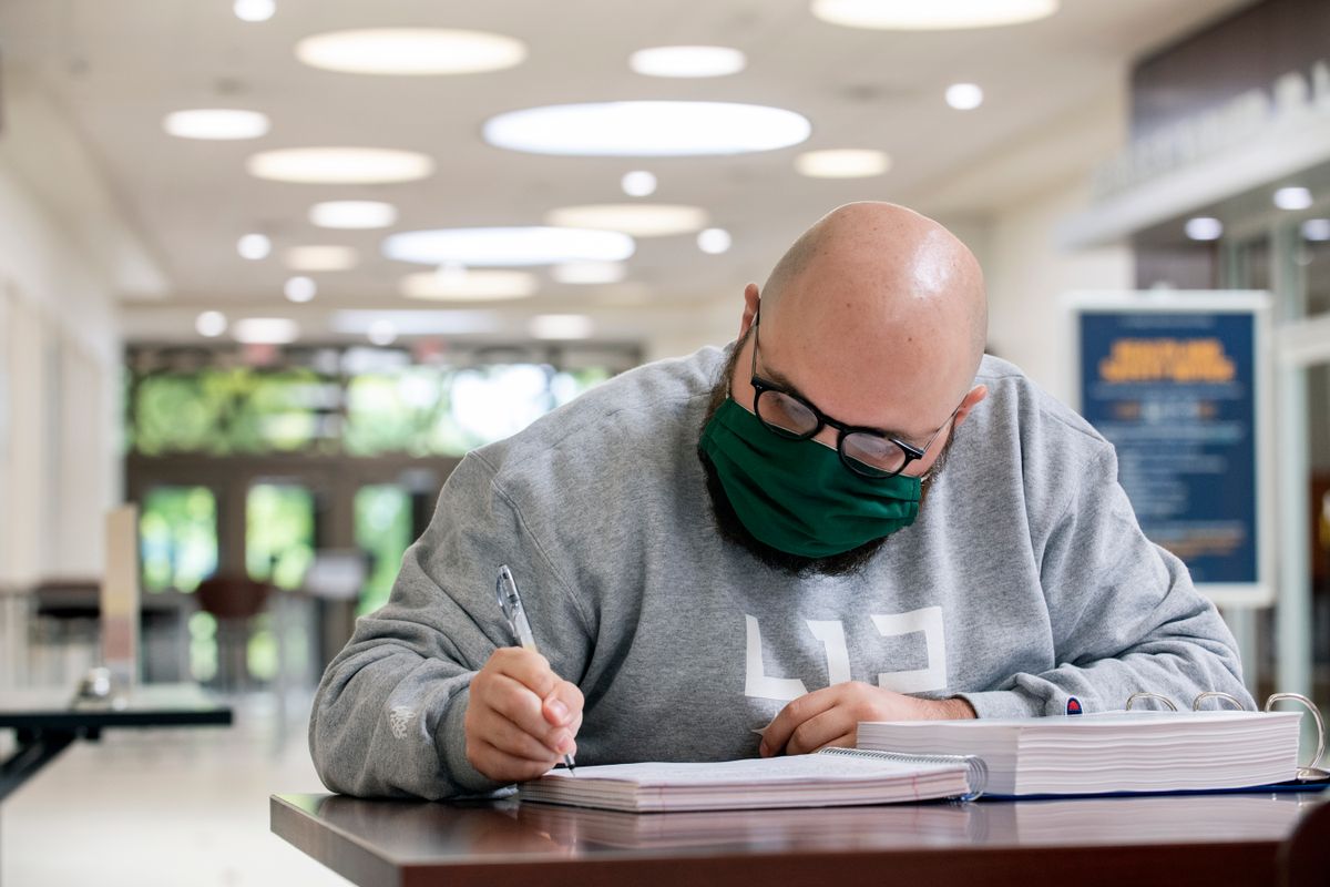 A bald man in black glasses, a mask and a gray sweatshirt with white lettering works through a stack of white papers while sitting at a brown desk