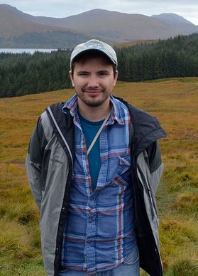 A person stands outside in front of mountains. He is wearing a plaid shirt, jacket and hat.
