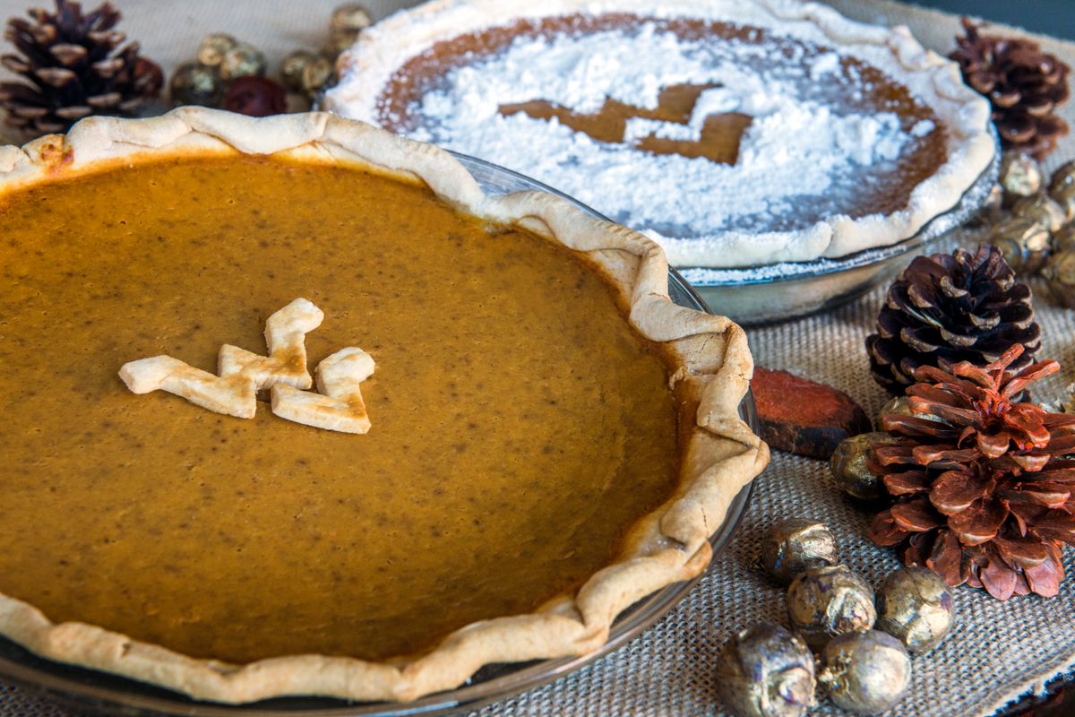 Photograph of two pumpkin pies, one with a Flying WV made out of crust dough placed in the center of the pie, and the other coated in powdered sugar minus a negative space Flying WV in the center. Both pies are sitting on a table decorated for the holiday