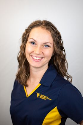 A woman with curly brown hair and blue eyes smiles with a blue and gold polo on