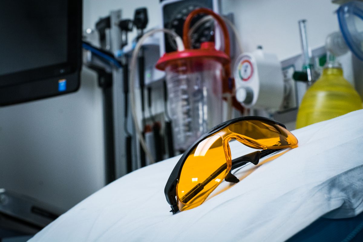 A pair of orange-colored glasses rests on a white surface with medical equipment in the background
