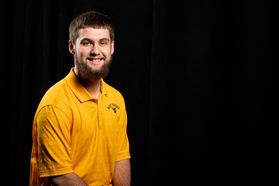 Photo of young man with dark hair and beard wearing gold shirt with flying WV below West Virginia against a dark background