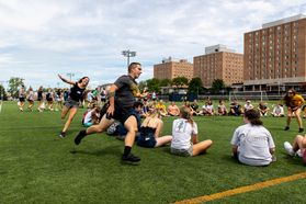 Students play games on the WVU Campus with the Towers residence halls in the background. The picture shows a female student chasing a male student around a circle of other students sitting on the grass. 