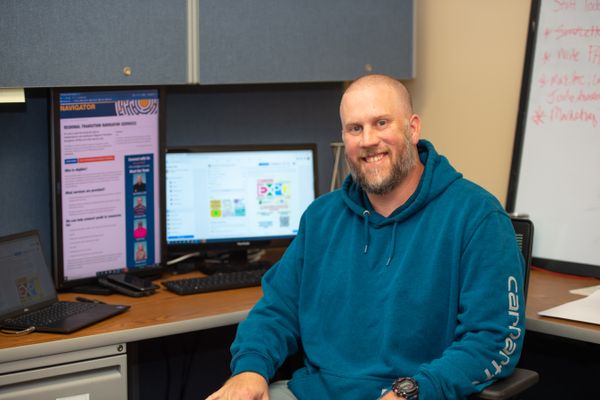 Regional Transition Navigator Program Manager Sam Wilkinson sits at a desk with computer monitors behind him. He is bald and is wearing a blue hooded sweatshirt.