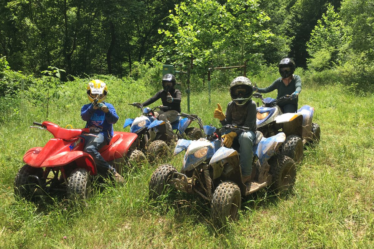 Four riders wearing helmets sit atop all-terrain vehicles in a grassy field
