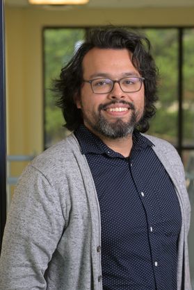 Headshot of WVU researcher Emmanuel Fonseca. He is standing in front of a window and is wearing a gray cardigan over a navy blue button-up shirt. He has shoulder length black hair and a salt and pepper goat tee. He also wears glasses. 