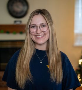 Headshot of WVU student Marissa Mangione. She is pictured inside with a beige wall behind her wearing a navy blue WVU T-shirt. She has long blonde hair and wears glasses. 