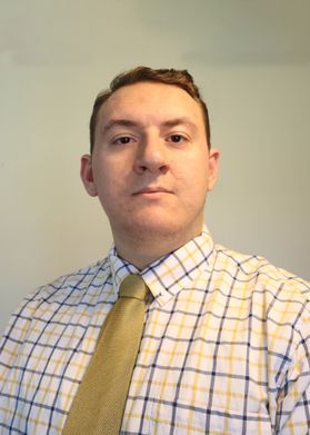Headshot of WVU student Jef Devlin. He is pictured inside with a white wall in the background. He is wearing a blue and yellow plaid dress shirt with a gold tie. He has short, brown hair. 