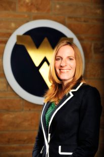 Blonde woman stands in front of flying WV logo in black and white suit jacket and green shirt