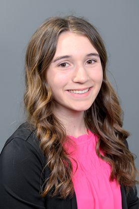 Headshot of WVU Bucklew Scholar Kierstn Posey. She is pictured against a gray background wearing a black jacket over a hot pink blouse. She has long, curly brown hair. 