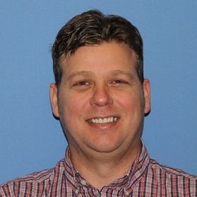Headshot of WVU professor Chad Pierskalla. He is pictured against a light blue background. He is wearing a red plaid shirt and has short brown hair. 