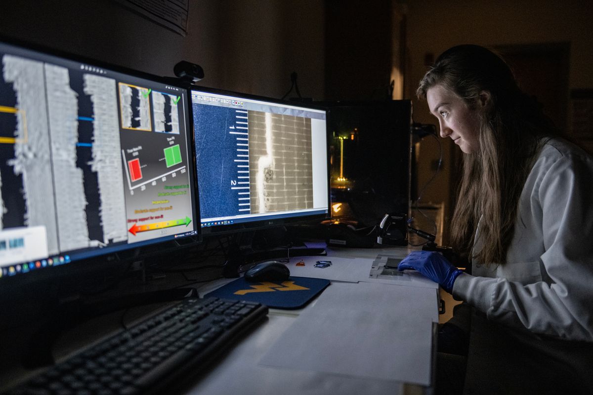 WVU student works in a lab. She is seated at a desk in a dark room and is looking at data on two computer screens. There are papers on the desk in front of her. 