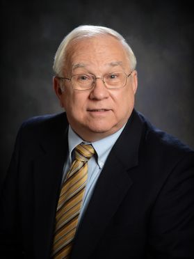 WVU honoree Robert Daily pictured in front of a dark gray background wearing a dark colored suit over a light colored dress shirt and striped tie. He has gray hair and wears glasses. 