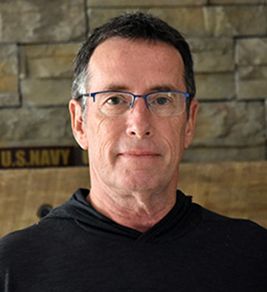 Headshot of sociology professor James Nolan. He's middle aged, wearing a black shirt and glasses and standing in front of a stone wall. 