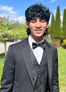 This is a portrait of Ayaan Shaik who is standing outside with trees in the background and wearing a tuxedo with a black patterned vest and tie.