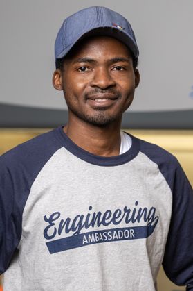 Headshot of WVU graduate student Raphael Oladokun. He is pictured wearing a white and gray baseball T-shirt with the words Engineering Ambassador on it. He is also wearing a blue baseball cap over his dark hair. 
