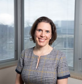 Headshot of WVU Cancer Center administrator Hannah Hazard-Jenkins. She is pictured standing in front of windows wearing a black, gray, gold and white plaid dress. She has short brown hair. 