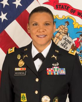 A headshot of a woman in a military uniform with the American flag behind her.