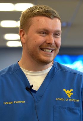 This is a picture of Carson Cochran, a dental surgery student who has short dark blond hair and is wearing blue scrubs over a white shirt with a flying WV and the words 'School of Dentistry' on his left chest.