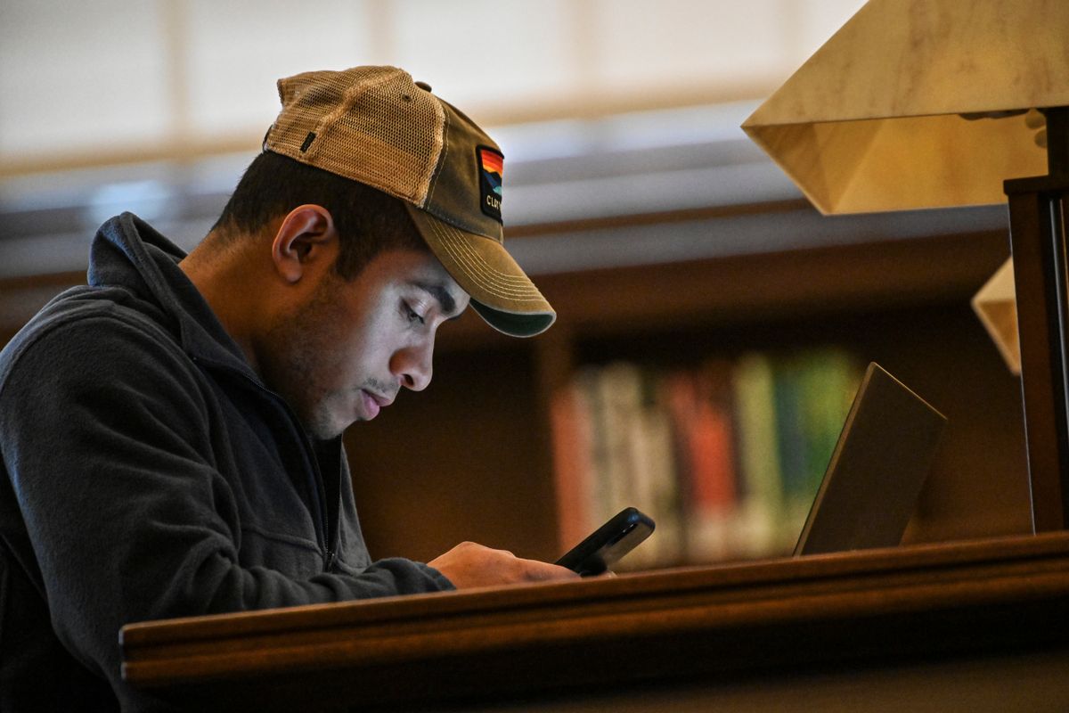 male student in hat looks at his cell phone in a dark library