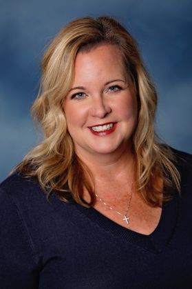 Headshot of WVU honoree Laurie Pollack. She is pictured in front of a light blue background and is wearing a navy blue V-neck sweater. She has long blonde hair and wears a cross necklace. 