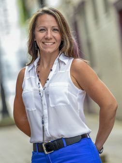 Headshot of WVU professor Nettie Freshour. She is standing outside wearing a sleeveless white top and royal blue pants cinched with a black belt. She has shoulder length blond hair and also has a long, dark beaded necklace around her neck. 