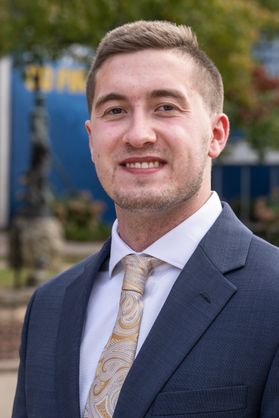 Headshot of WVU student Thomas Williams. He is pictured outside wearing a navy blue sport coat over a white dress shirt with a gold, paisley tie. He has short, light colored hair. 