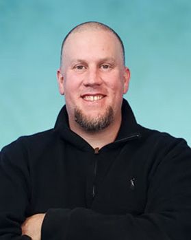 Headshot of WVU researcher Sam Wilkinson. He is pictured against a light blue background and has his arms crossed. He is wearing a black 1/4 zip sweater, has a shaved head and a goat tee. 