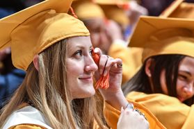 A graduate with long blonde hair and wearing a gold cap and gown moves an orange tassle to signify graduating from WVU.