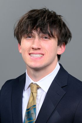 Headshot of WVU Bucklew Scholar Benjamin Golden. He is pictured against a light gray background and is wearing a navy blue suit with a white dress shirt and patterned tie. He has medium length brown hair. 