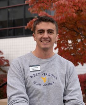 Headshot of WVU student Daniel Givler. He is pictured outside with a tree in the background full of red leaves. He is wearing a WVU branded gray sweatshirt and has a name tag on. He has short brown hair. 