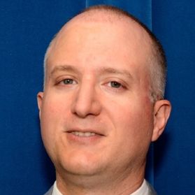 Headshot of WVU professor Edward Timmons. He is pictured against a blue background and only his head is visible in the frame. He is balding with gray hair around the sides of his head. He is smiling slighting in the photo. A white dress shirt is visible. 