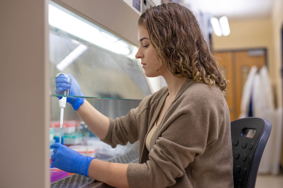 A research sits at a long counter with flourescent lighting shining down. The research has shoulder length light brown hair, is wearing a brown sweater and has on blue latex gloves while working with lab equipment.