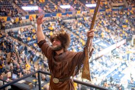 At the Coliseum, Mikel Hager participates in the cheeroff competition. Hager is wearing the buckskins with a gold belt and coonskin cap. Both of his arms are raised. The rifle is in his left hand.