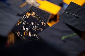 An embroidered graduation cap is shown among others. It reads, "We rise by lifting Others." The OT is gold. The rest of the words are in white letters.