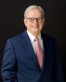 A man with short white hair and round black glasses. He is wearing a white button up dress shirt with a navy blue suite jacket and a light pink tie
