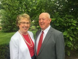 Woman wearing red shirt, white sweater and pearls standing next to man wearing grey suit with red tie in front of green grass and trees