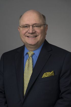 Headshot of WVU honoree J. Wayne Richards. He is pictured against a gray background wearing a dark colored suit, blue dress shirt and matching yellow and blue tie and pocket square. He wears glasses and has gray hair. 